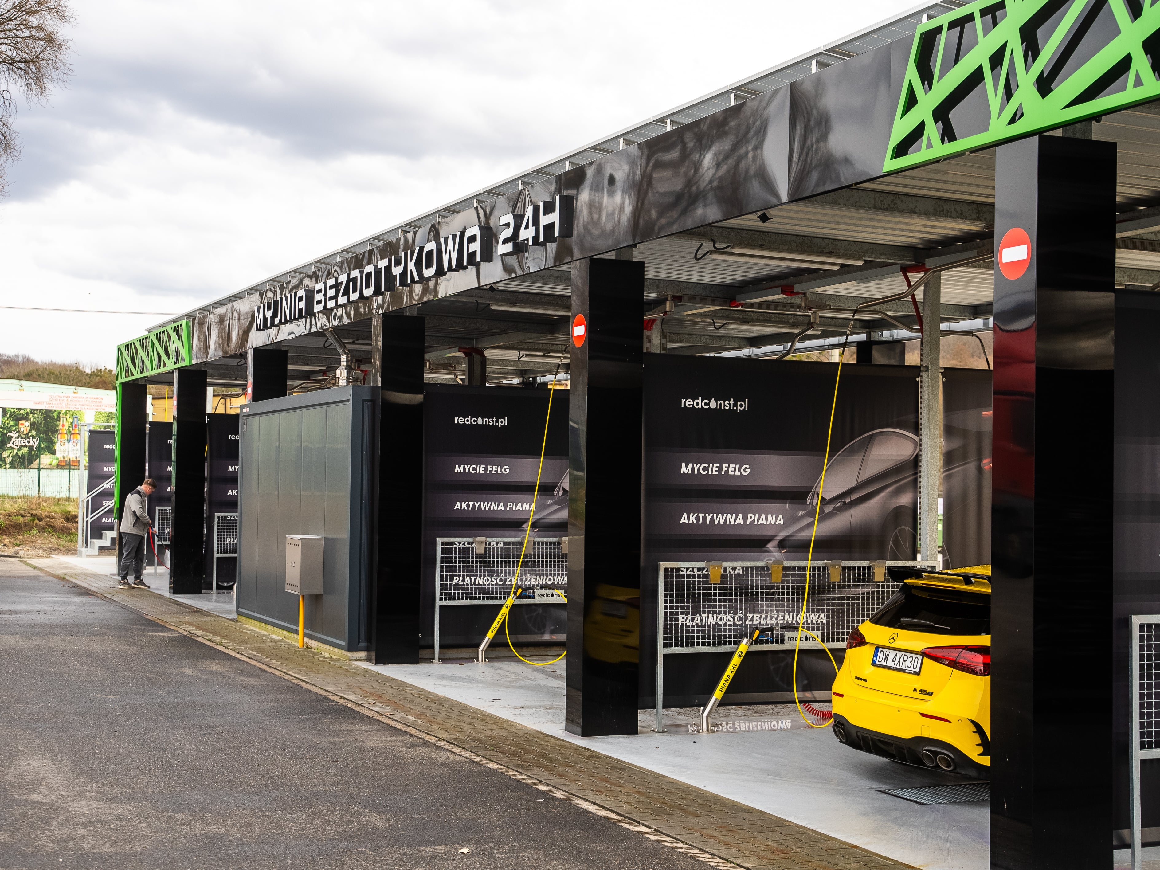 Wnętrze myjni bezdotykowej 24H, z widokiem na stanowiska do mycia samochodów. W tle widoczne informacje o usługach, takich jak mycie felg i aktywna piana, a także żółty samochód czekający na mycie. Idealne dla miłośników czystości pojazdów, dostawca nowoczesnych rozwiązań w branży myjni.